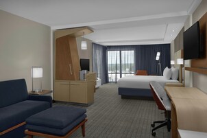 Habitación de hotel con cama, escritorio, sofá, televisor y un amplio ventanal con cortinas.