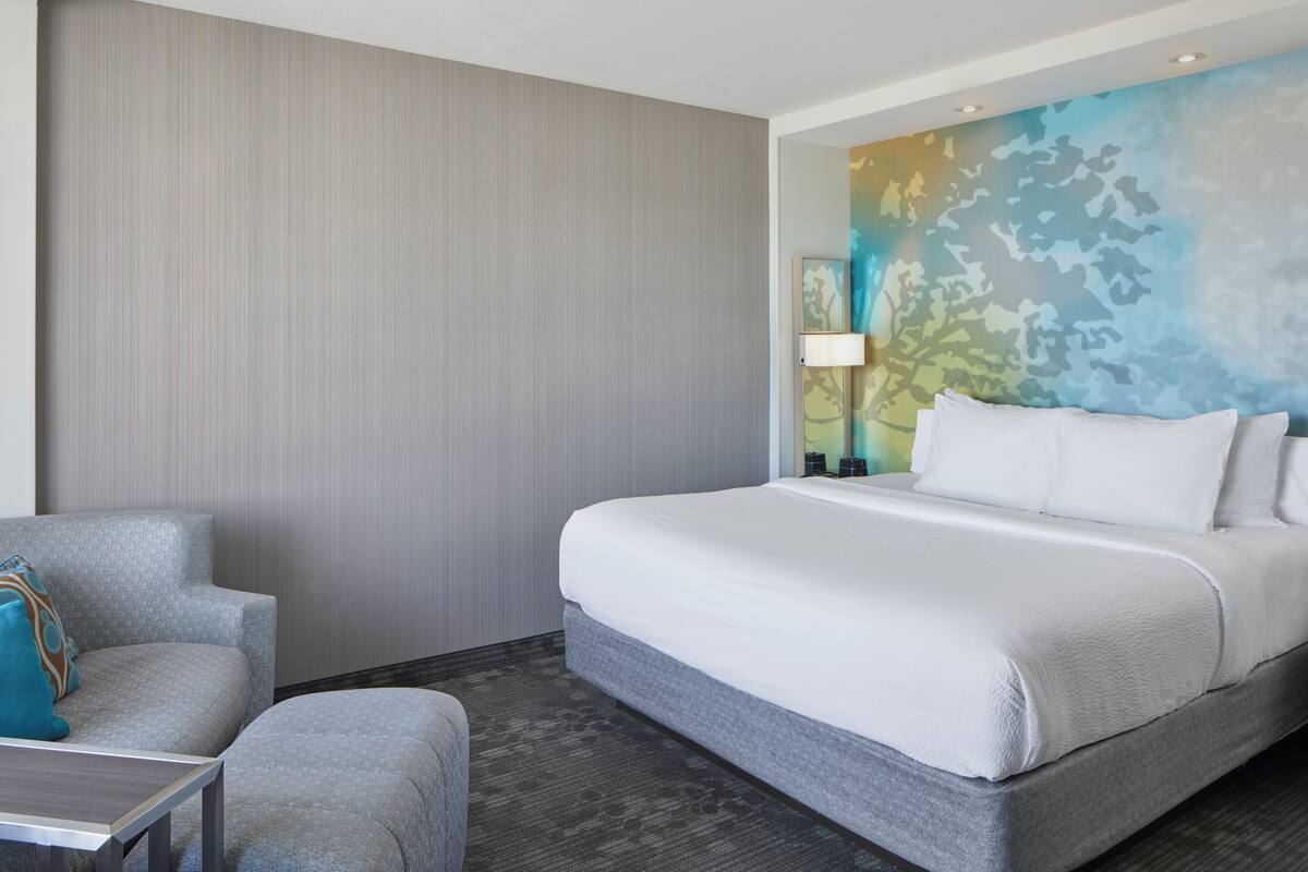A hotel room with a bed, a chair, a table, and a wall with a painting.