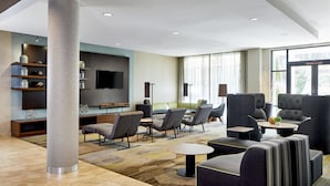 Un moderno vestíbulo de hotel con un pilar grande, zona de estar con sofás y sillones, un televisor de pantalla plana en la pared y un amplio ventanal que proporciona luz natural.