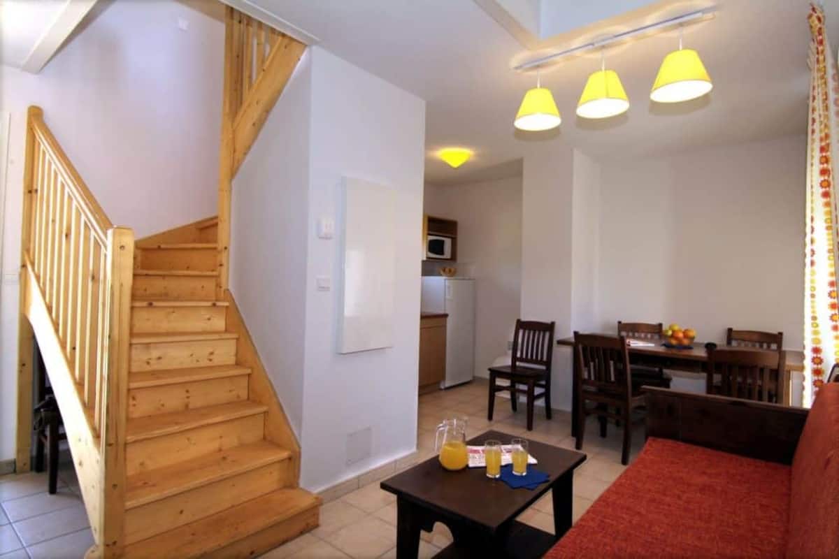 Un escalier menant à un deuxième étage, un coin repas avec des chaises et une table en bois, et une cuisine à l’arrière-plan.