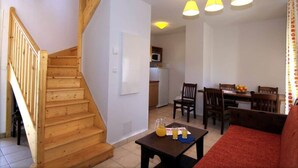 Una escalera conduce a un segundo piso, un comedor con sillas de madera y una mesa, y una cocina al fondo.