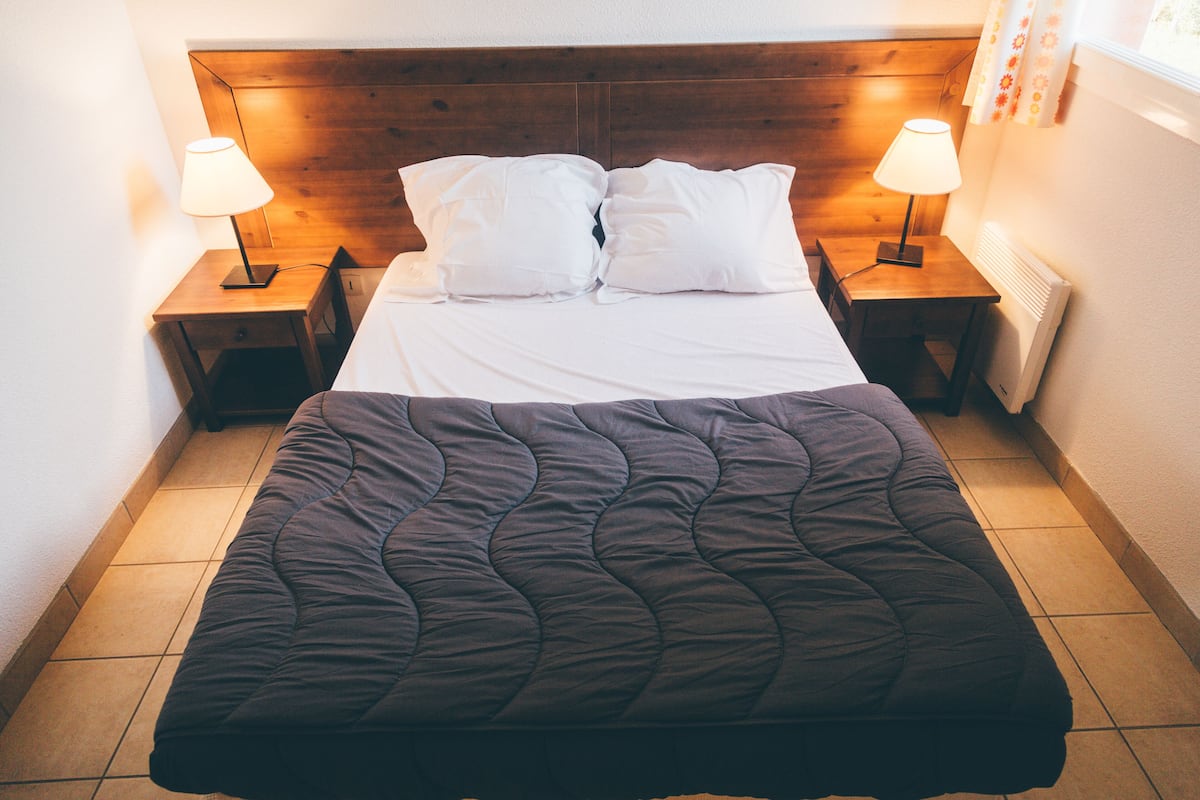 Un lit bien fait, recouvert d’une couverture sombre, deux tables de chevet avec des lampes et une tête de lit en bois.