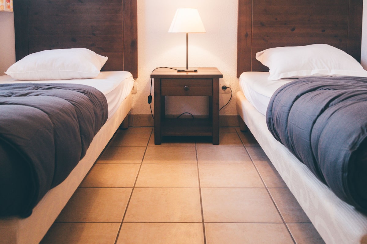 Une chambre d’hôtel avec deux lits simples, une table de chevet avec une lampe et un sol carrelé.