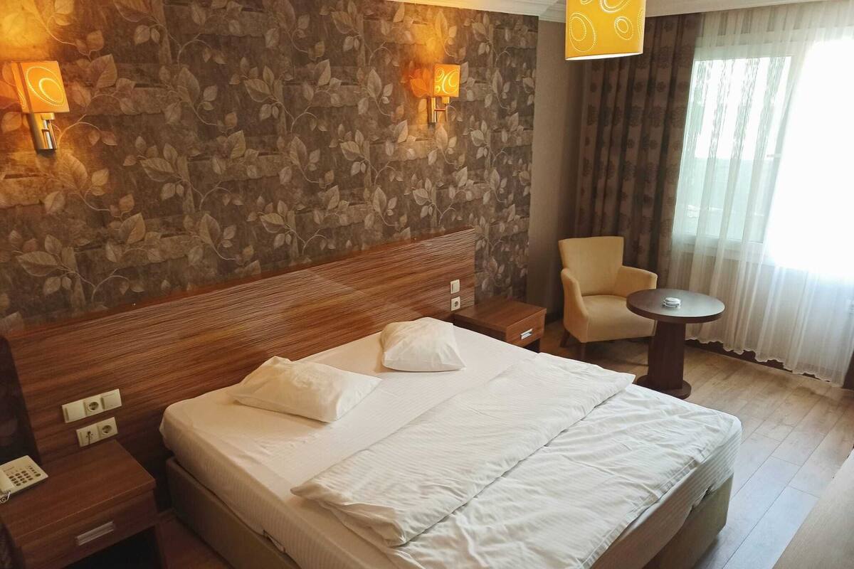 A hotel room with a large bed, a wooden headboard, two bedside tables, a chair, a small round table, and a floral wallpaper.