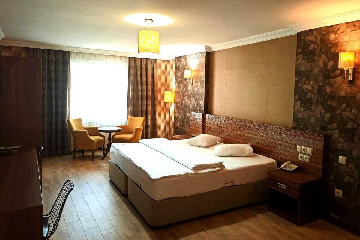 A hotel room with a large bed, a desk with a telephone, two chairs, a small table, and a window with curtains.