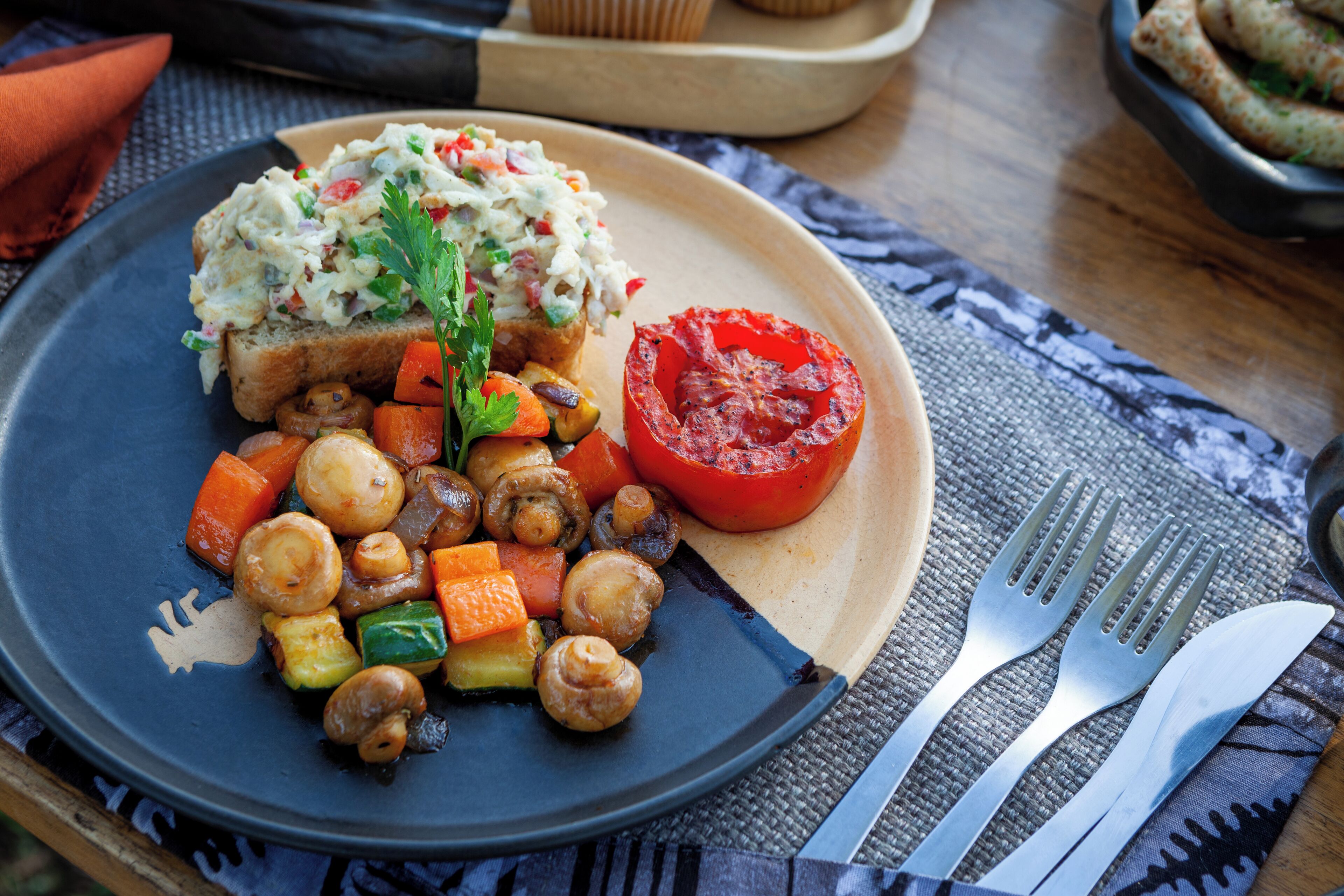 A plate with roasted vegetables, a slice of tomato, and a piece of bread with a creamy topping.