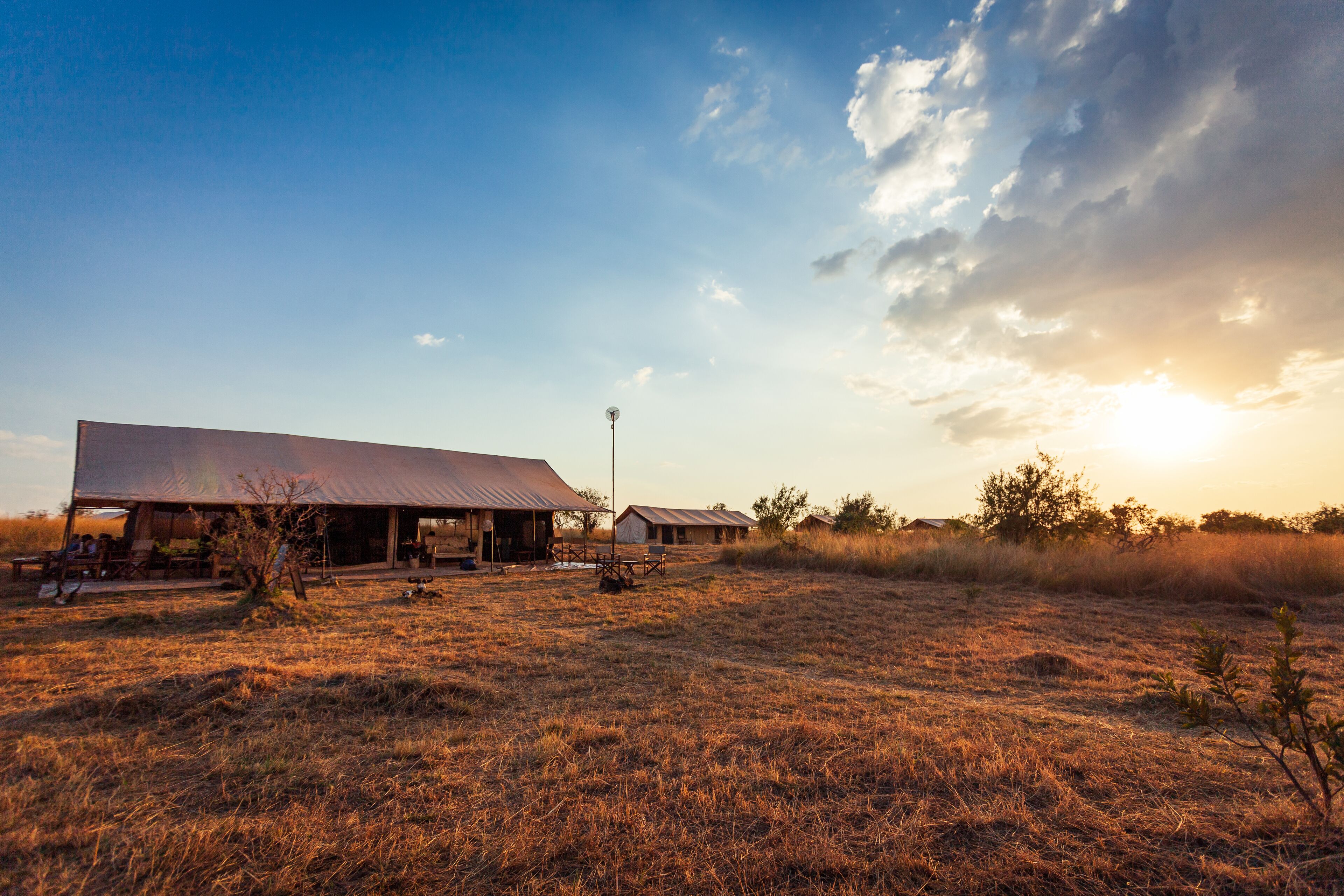 A large barn with a flat roof and a small adjacent building, surrounded by dry grass fields and a clear sky.