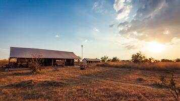 A large barn with a flat roof and a small adjacent building, surrounded by dry grass fields and a clear sky.