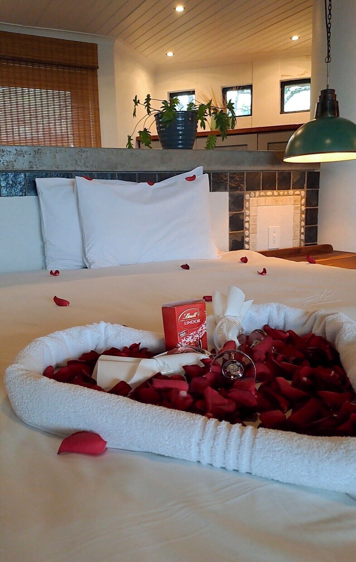 A neatly made bed with rose petals, a bottle of Lindor chocolates, and a white bathrobe.