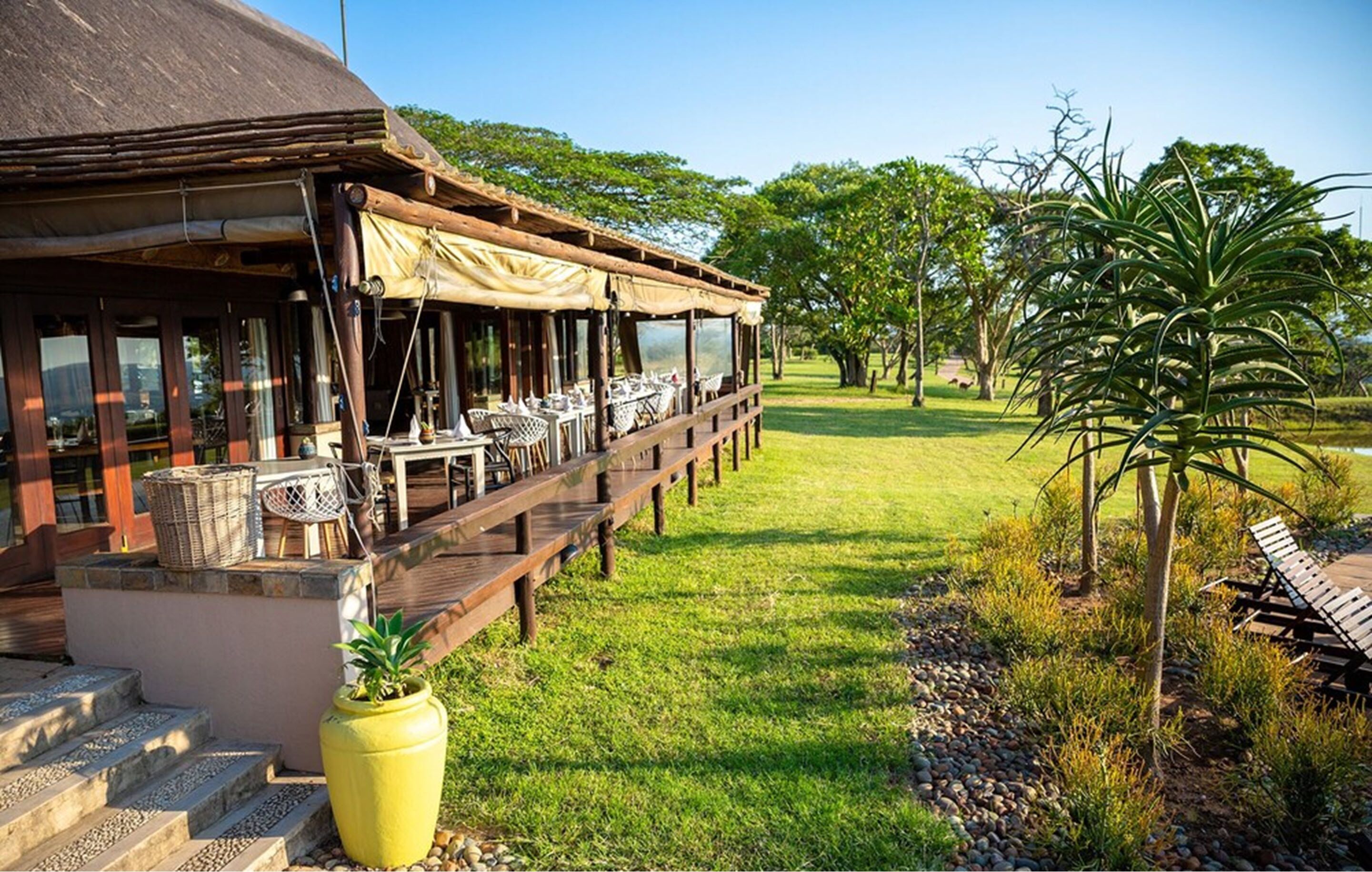 A restaurant with outdoor seating, a thatched roof, and a view of a grassy area with trees.
