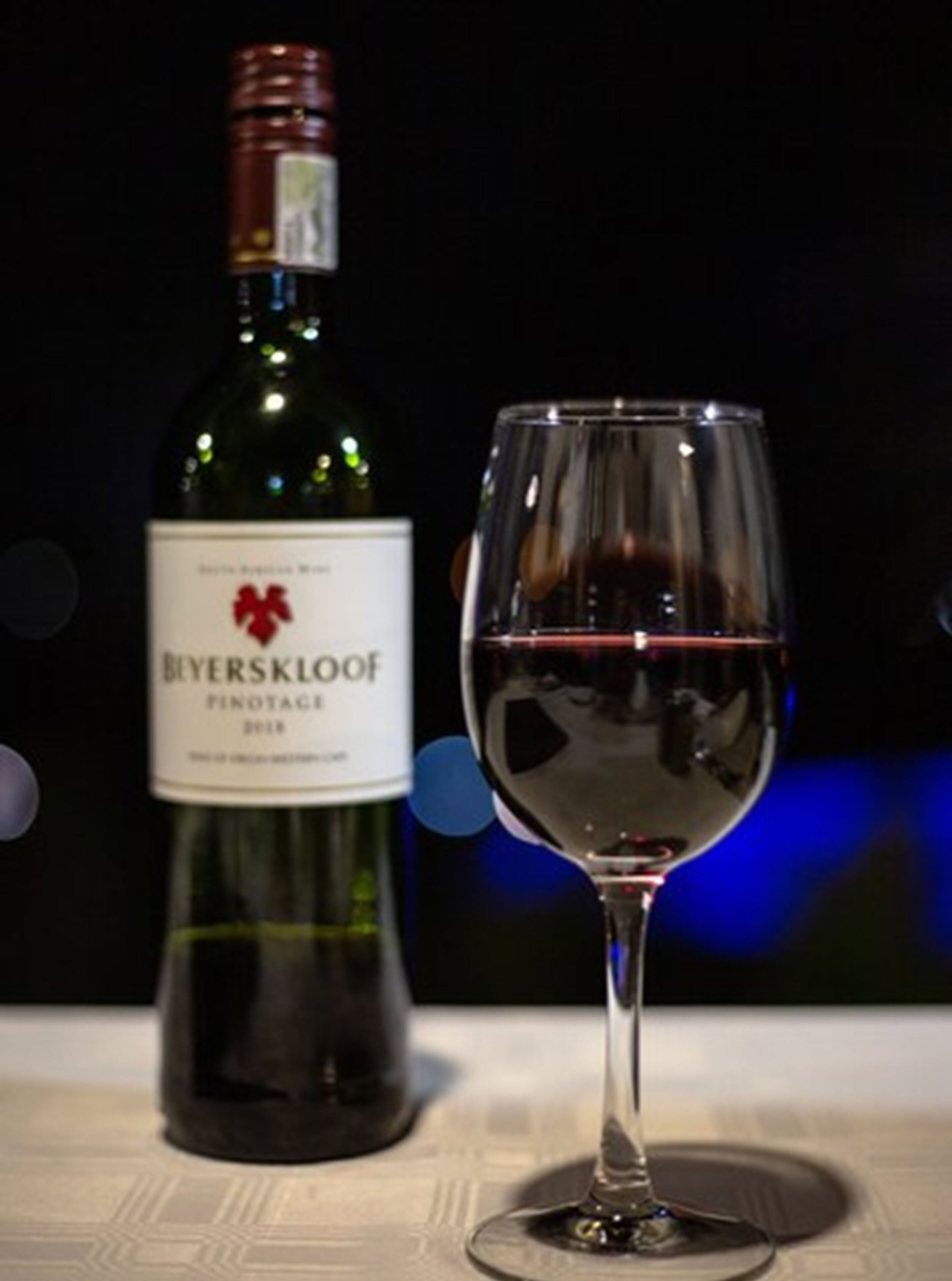 Uma garrafa de vinho Beyerskloof Pinotage 2018 e uma taça de vinho vazia sobre uma mesa.