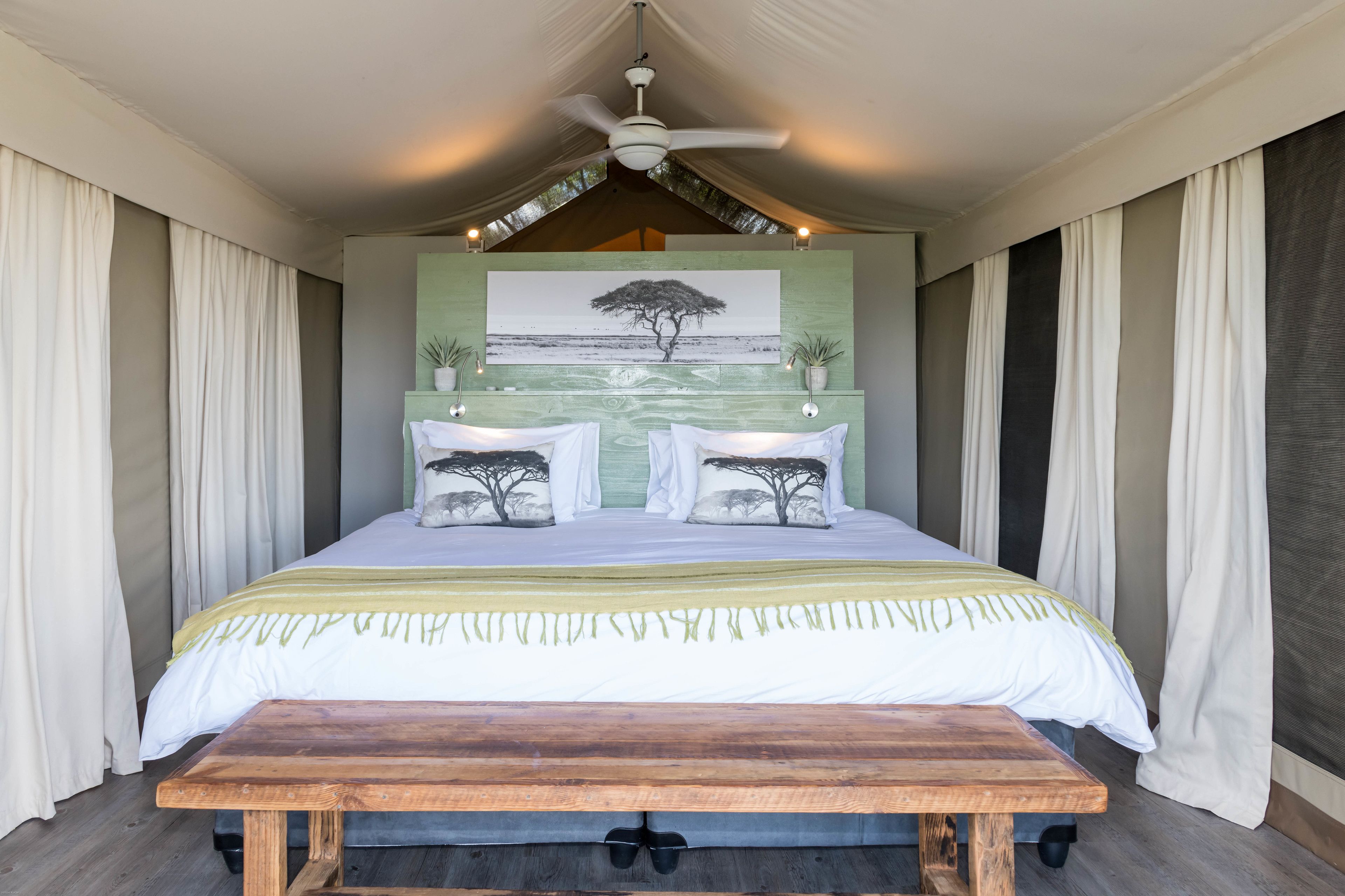 Une chambre à coucher avec un lit, un banc en bois, un ventilateur de plafond et un tableau représentant un arbre.