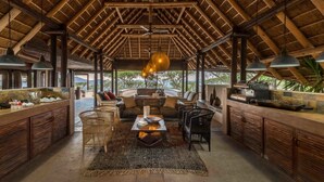 Un interior rústico con techo de madera alto, una zona de estar central con una mesa y una barra con una parrilla.