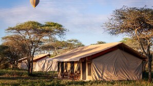 A safari tent with a table and chairs set up for dining, surrounded by trees. - Gnu Ndutu Camp (Ngorongoro)
