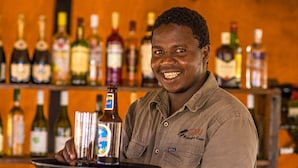 A person holding a tray with a bottle and glasses, standing in front of a bar with various bottles and glasses on shelves. - Gnu Ndutu Camp (Ngorongoro)