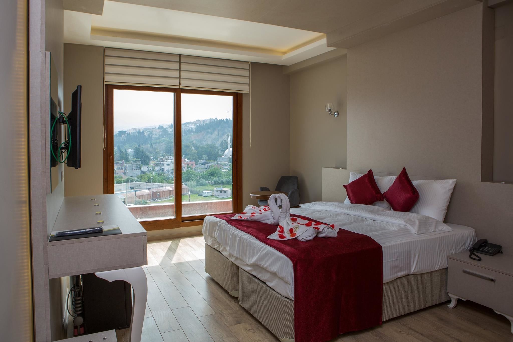 A hotel room with a bed, bedside table, and a desk with a chair. There is a large window with a view of a cityscape.
