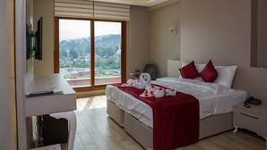 A hotel room with a bed, bedside table, and a desk with a chair. There is a large window with a view of a cityscape.