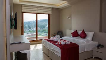 A hotel room with a bed, bedside table, and a desk with a chair. There is a large window with a view of a cityscape.