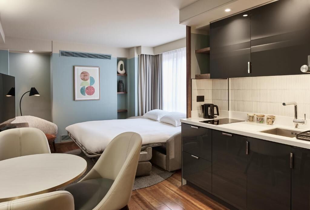 A modern hotel room with a bed, a small dining table, a sofa, a kitchenette, and a large window with curtains.