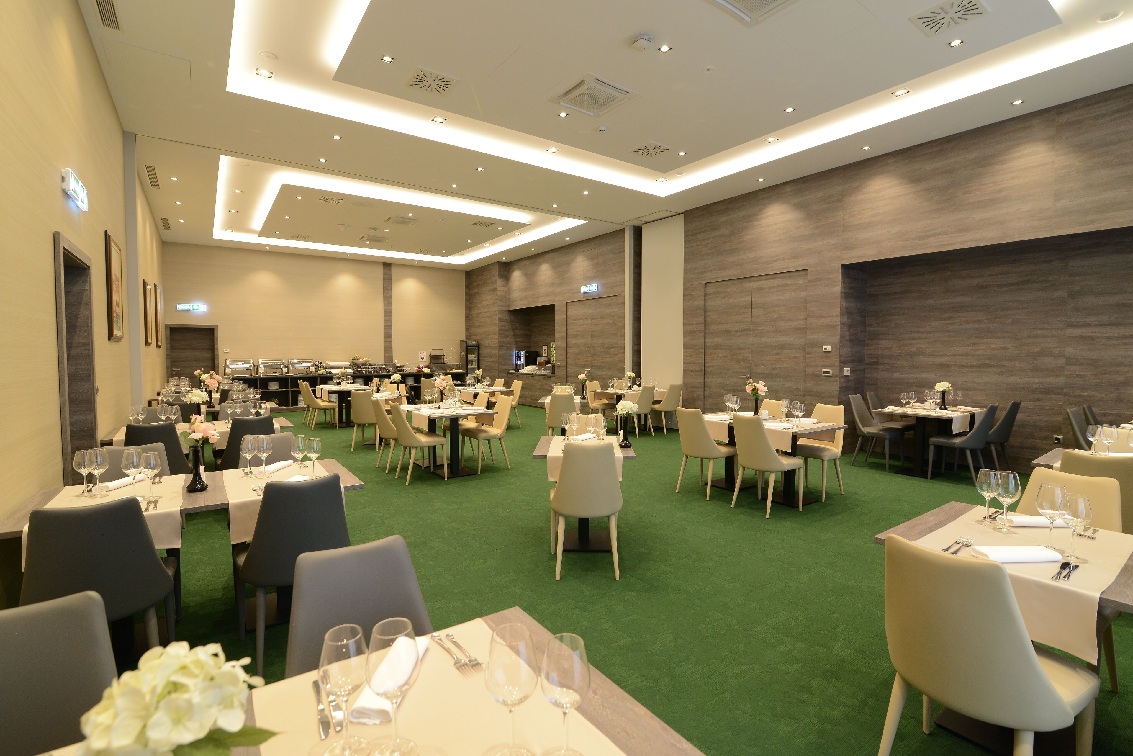 A spacious hotel dining area with multiple tables and chairs, green carpet, and modern lighting.