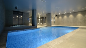 Una piscina interior con suelo y paredes de azulejos, rodeada por un diseño moderno con iluminación empotrada y un panel de vidrio.