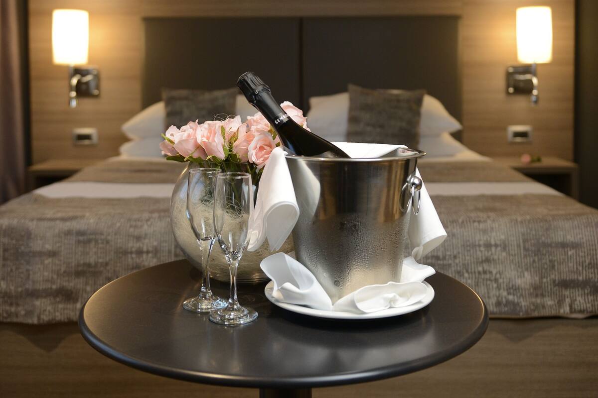 A hotel room with a bed, bedside lamps, a table with a champagne bottle, glasses, and a flower arrangement.