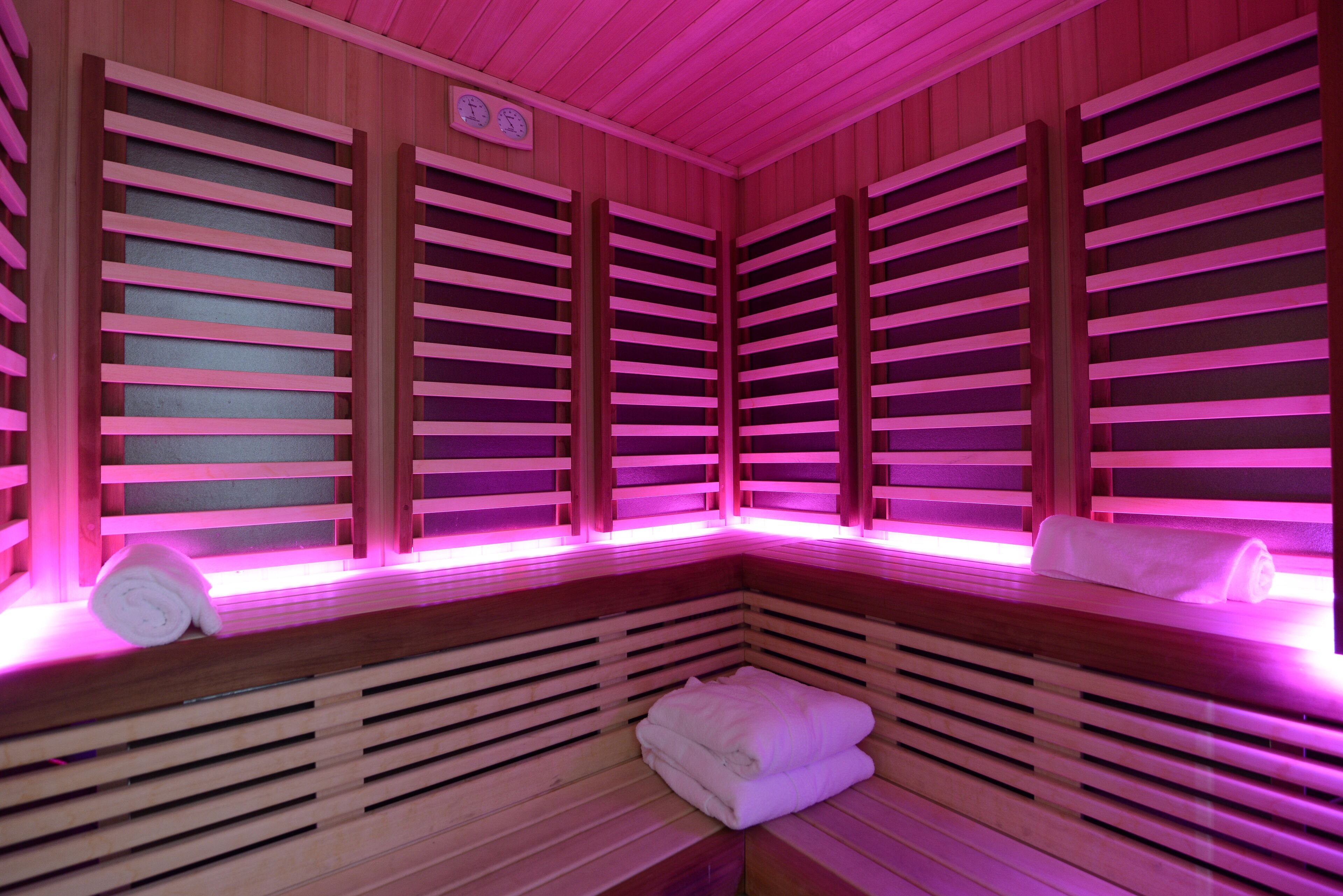 A sauna with wooden benches and slatted walls, pink lighting, and towels on the benches.