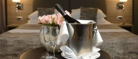 Habitación de hotel con cama, lámparas de noche, una mesa con una botella de champán, copas y un arreglo floral.