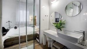 A modern hotel room with a glass-enclosed shower, a white bed, a large mirror, and a white sink.