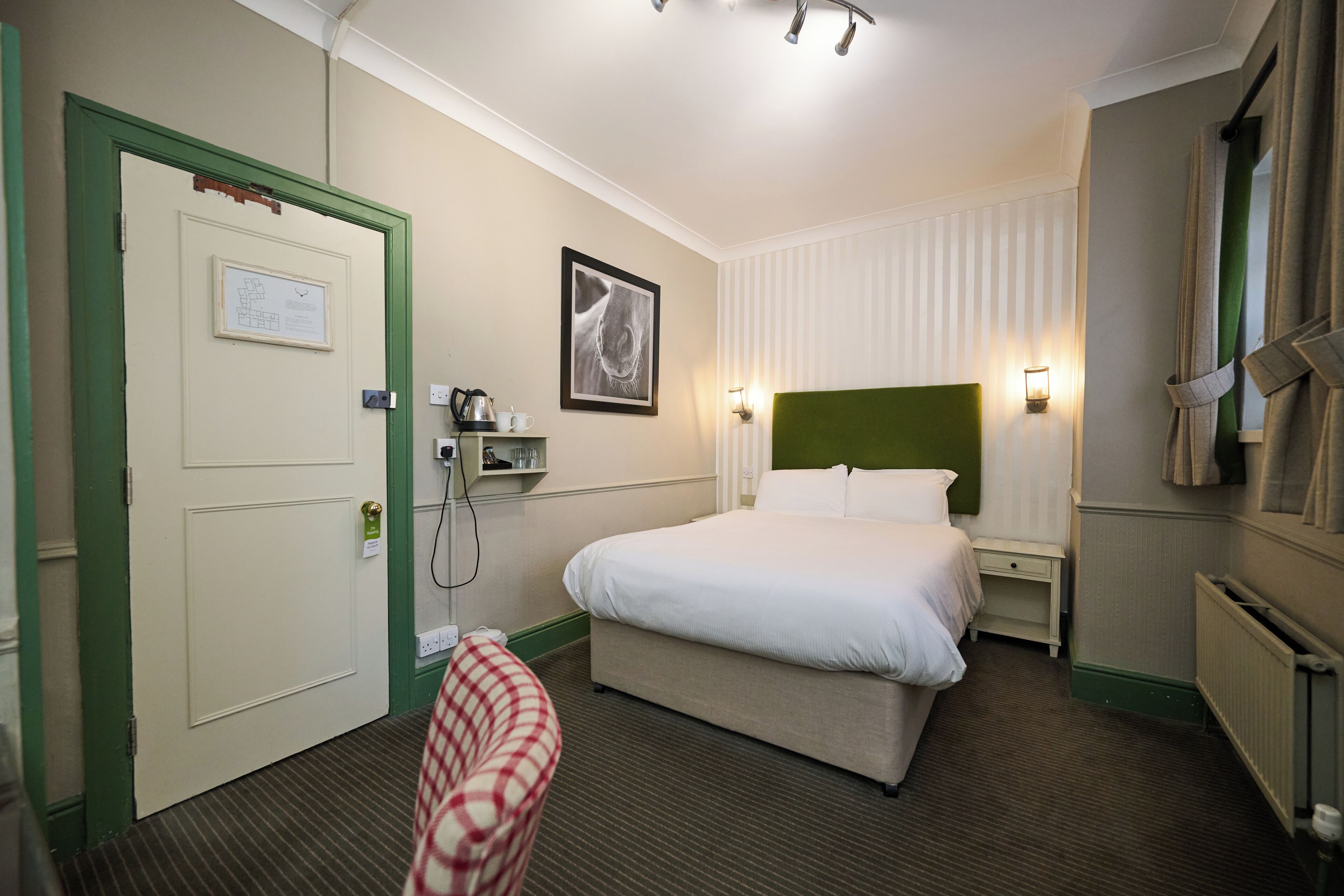 Una camera d'albergo con un letto, una sedia, un comodino, una mensola a parete e un termosifone.
