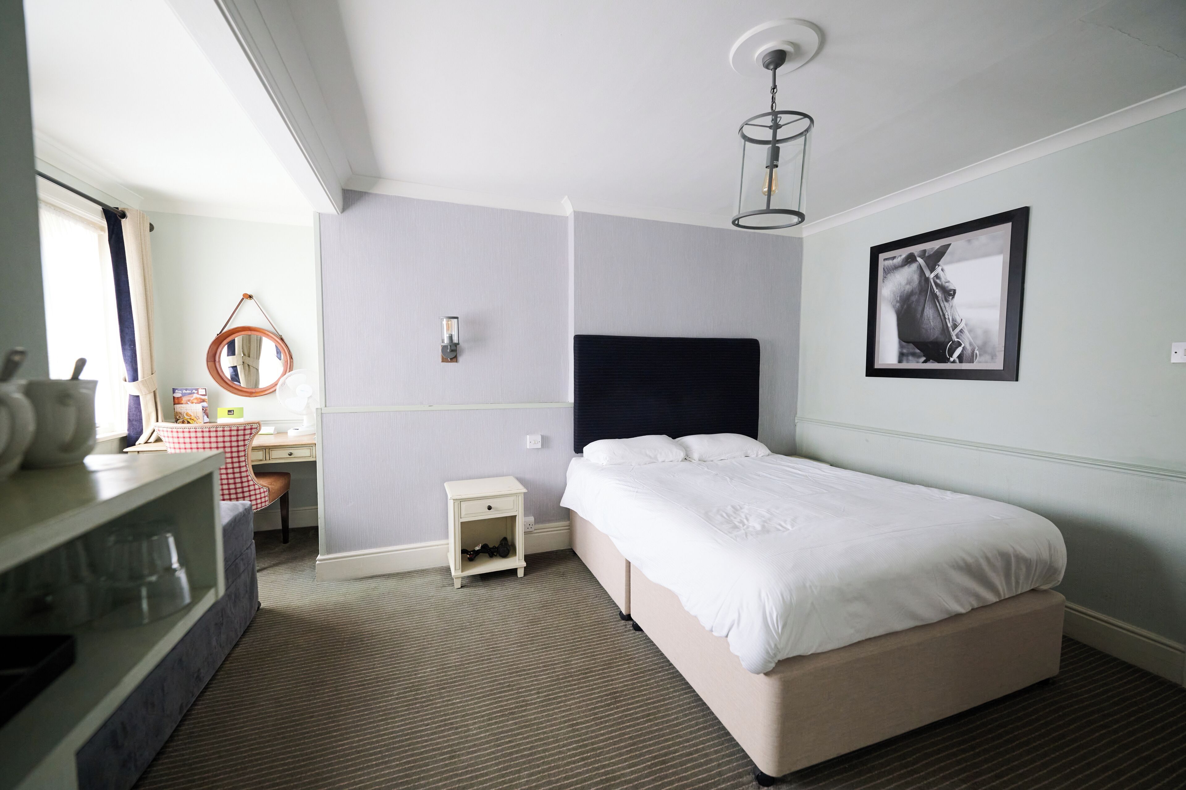 A hotel room with a bed, a desk with a chair, a small bedside table, a mirror, and a framed picture on the wall.