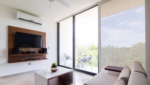 A modern living room with a flat-screen TV, a sofa, a coffee table, and a large window offering a view of greenery.