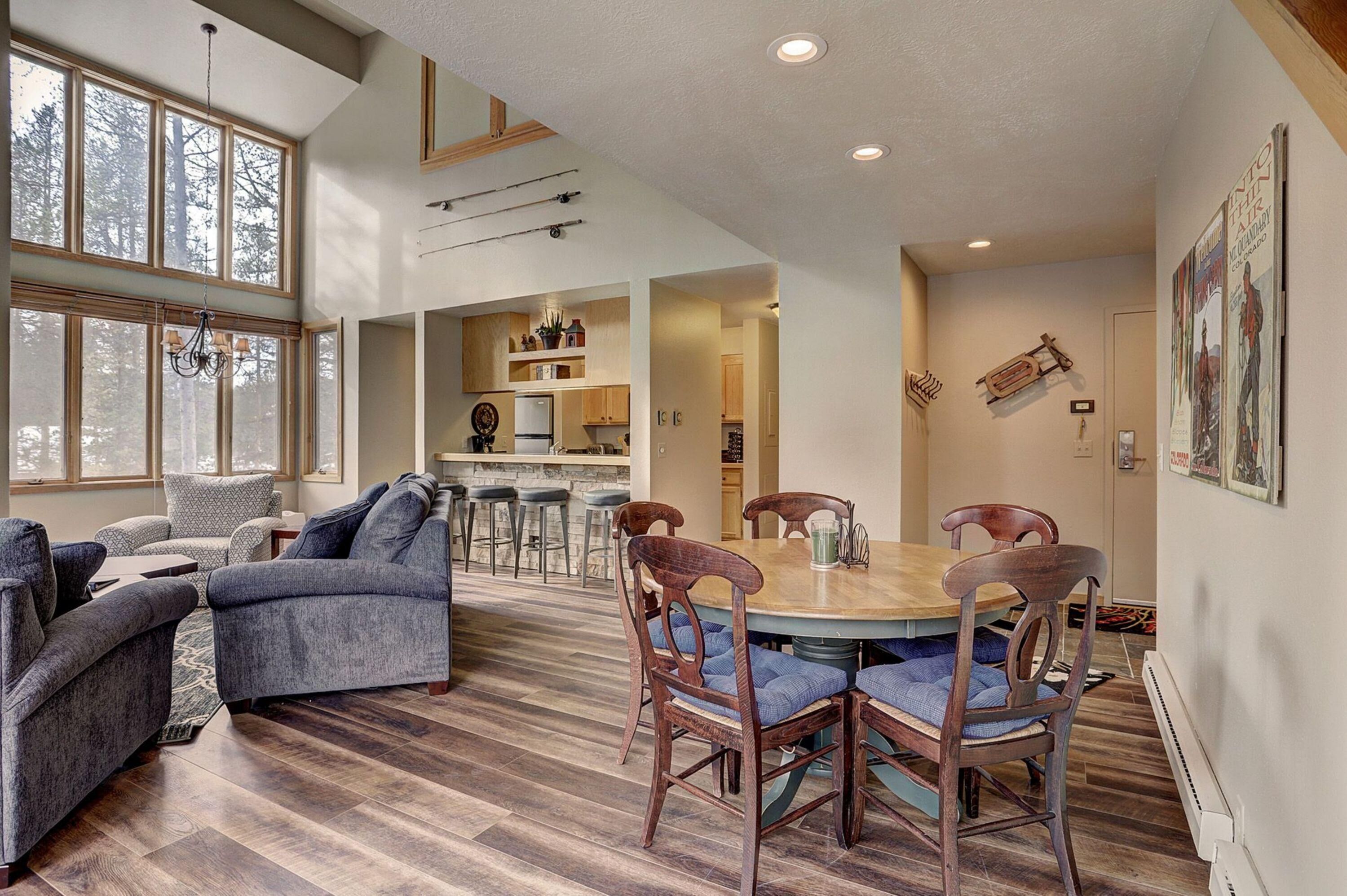 A spacious living area with a large window, a dining table with chairs, and a kitchen in the background.