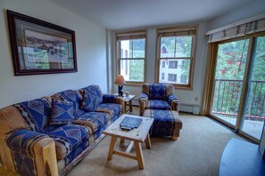 A living room with a patterned sofa, a small wooden table, and two armchairs. There is a framed picture on the wall and a window with a view of houses and trees. - Silver Mill 8227 (Keystone)