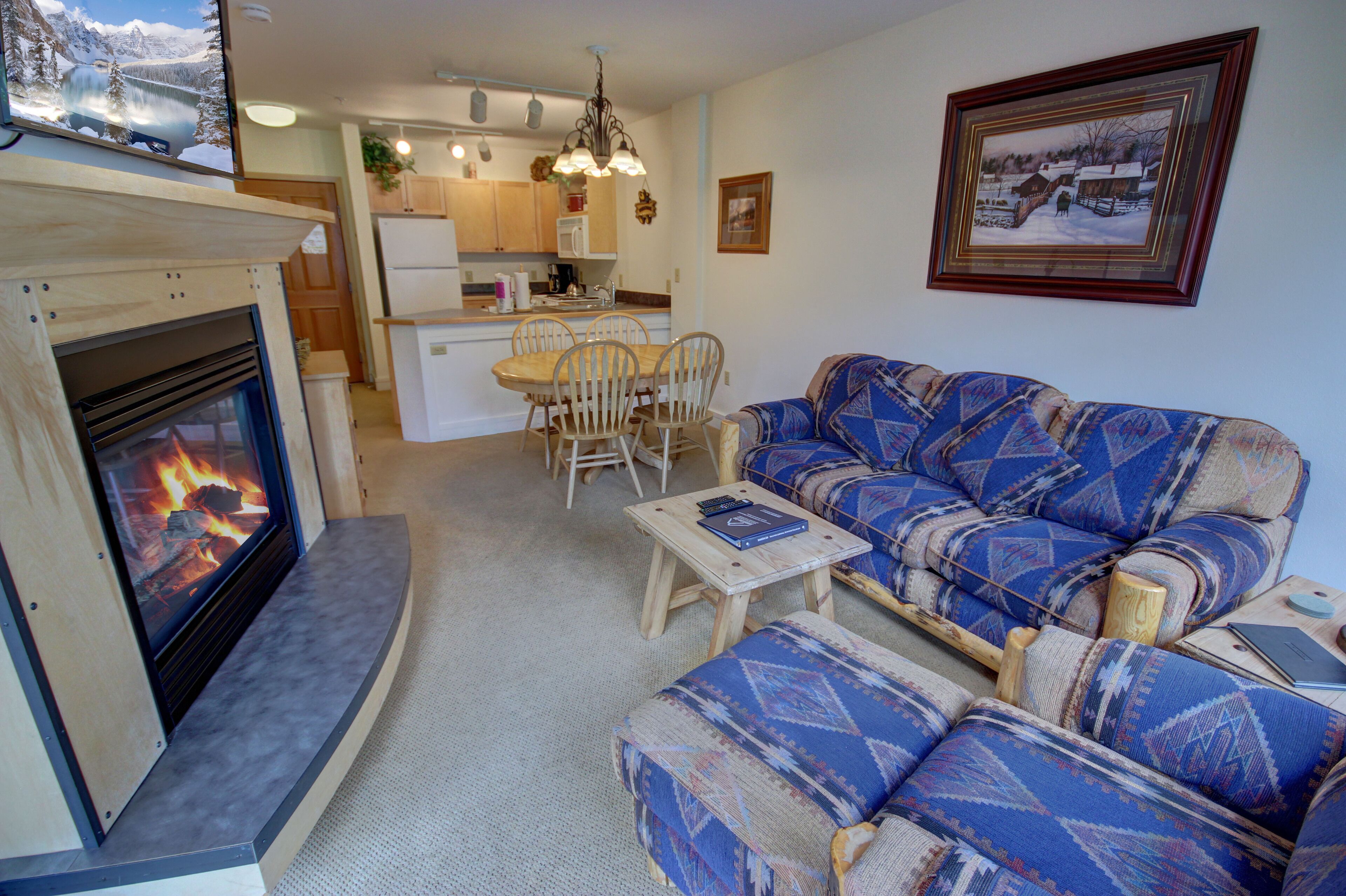 A cozy living room with a fireplace, a patterned sofa, a dining table with chairs, and a kitchen in the background.