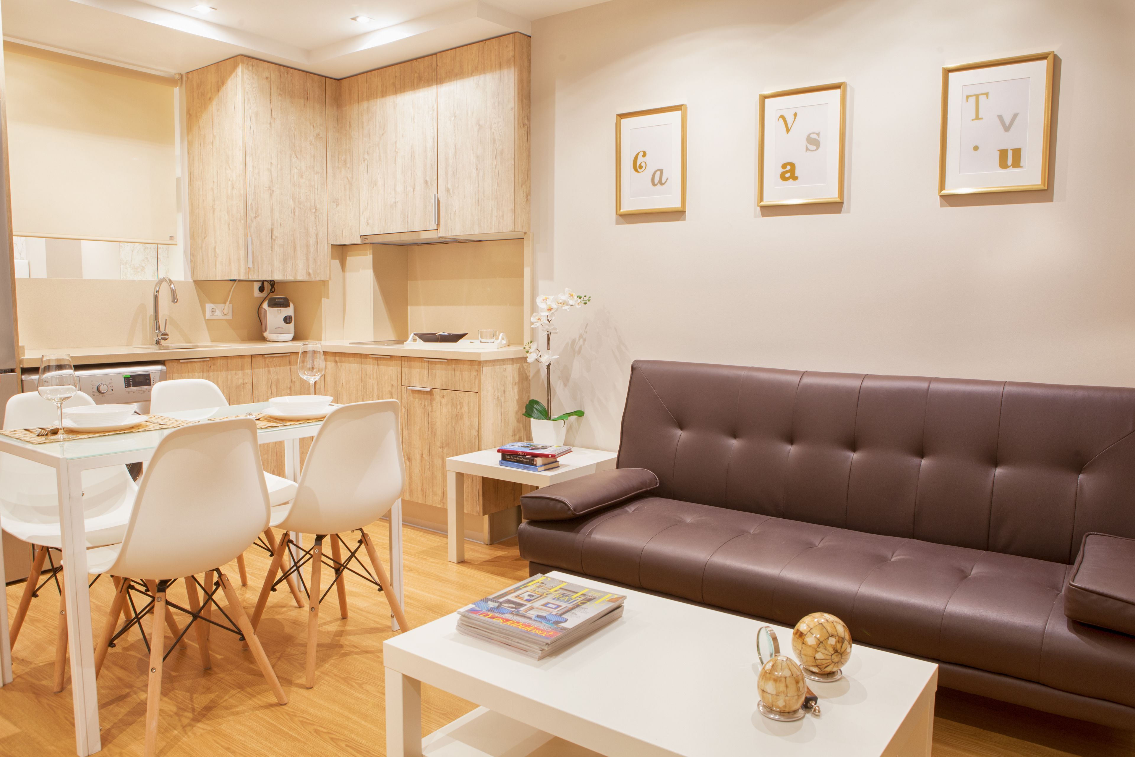 A modern living room with a brown leather sofa, a dining area with white chairs and a glass table, and a kitchen with wooden cabinets.