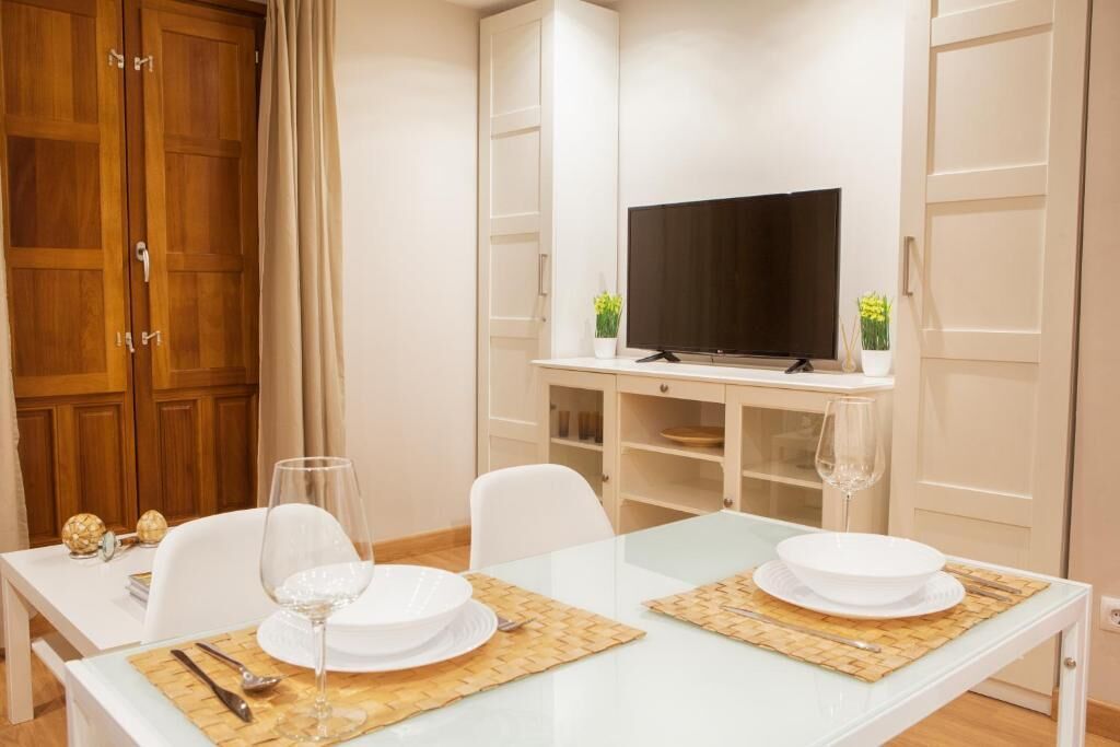 A dining area with a glass table, white chairs, and a television on a shelf.