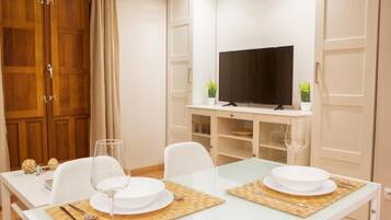 A dining area with a glass table, white chairs, and a television on a shelf.
