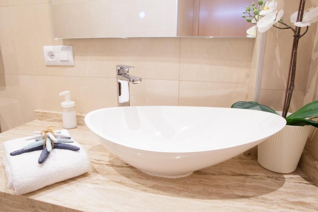 A bathroom with a large white vessel sink, a towel with decorative items, a soap dispenser, and a potted plant.