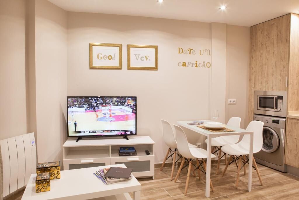 A modern living room with a flat-screen TV, white furniture, and a kitchen area with a microwave and washing machine.