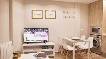 A modern living room with a flat-screen TV, white furniture, and a kitchen area with a microwave and washing machine.