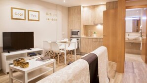 Una sala moderna con un televisor de pantalla plana, un comedor con mesa y sillas, una cocina con armarios de madera y electrodomésticos, y un baño visible a través de una puerta abierta.