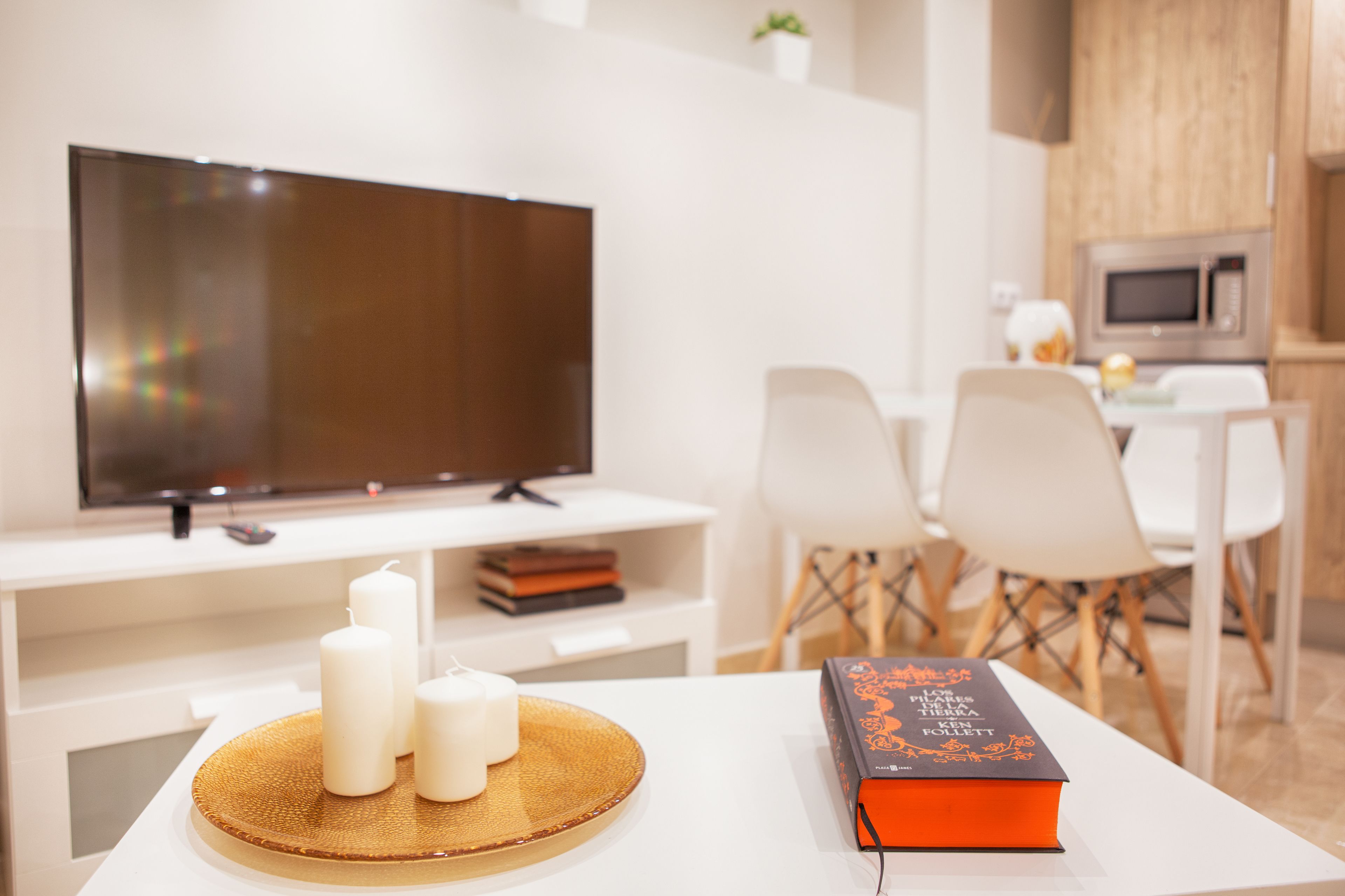A modern living room with a flat-screen TV, a white table with candles and a book, and a dining area with white chairs and a small table.