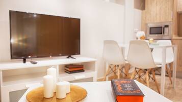 A modern living room with a flat-screen TV, a white table with candles and a book, and a dining area with white chairs and a small table.