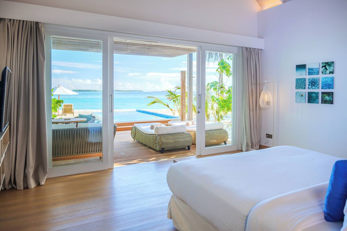 Une chambre d’hôtel dotée d’un grand lit, d’une porte-fenêtre coulissante donnant sur un balcon avec vue sur la mer, d’un petit coin salon avec une table et des chaises, et d’un mur orné de tableaux.