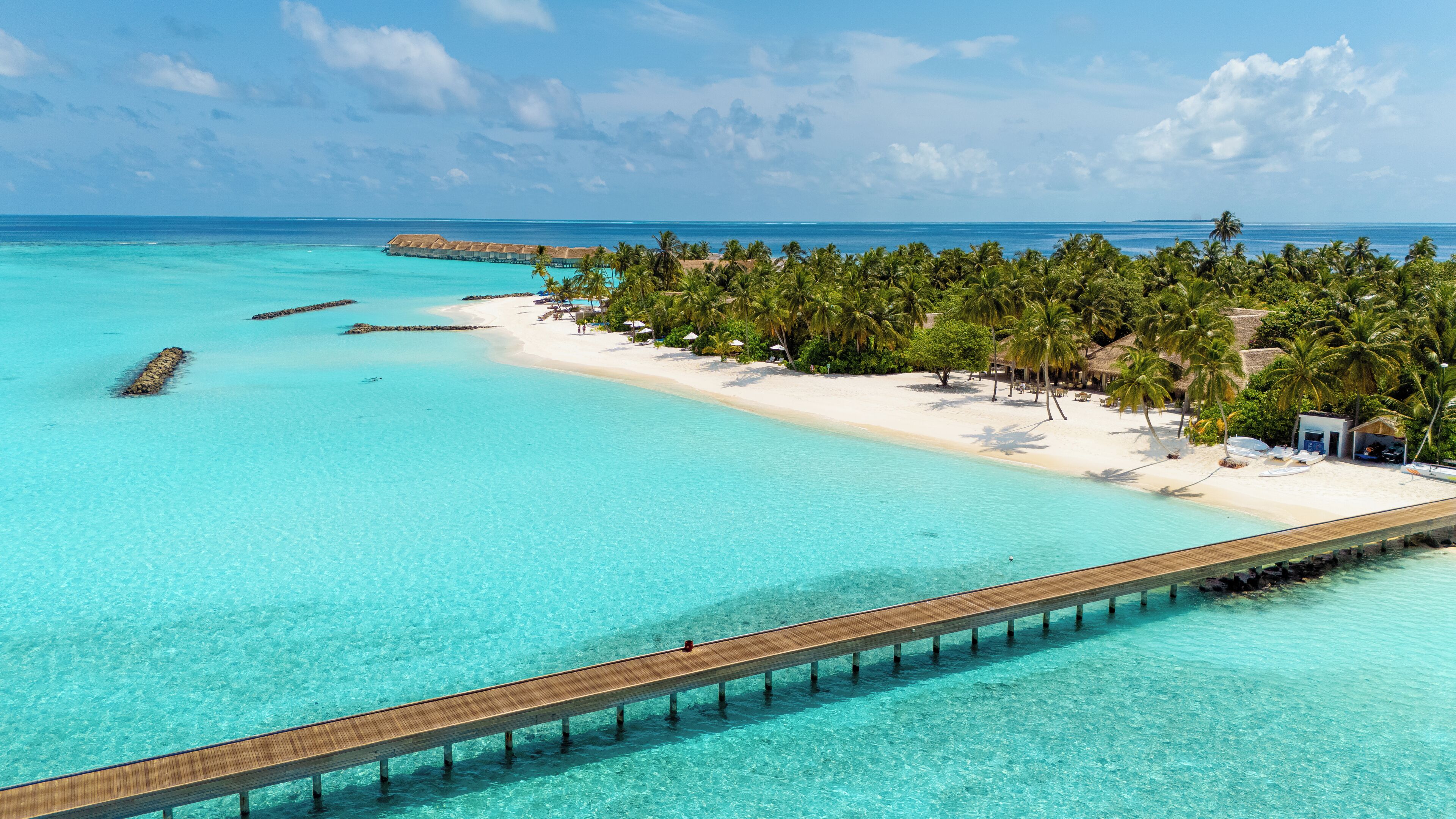 Un complexe hôtelier de bord de mer doté d’une jetée en bois s’avançant dans des eaux turquoise, d’une plage de sable et de palmiers.