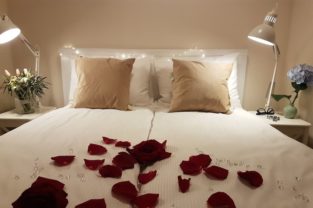 Una cama cuidadosamente tendida adornada con pétalos de rosa y una sola rosa, dos almohadas color beige, y una mesita de noche con una lámpara y un jarrón de flores.