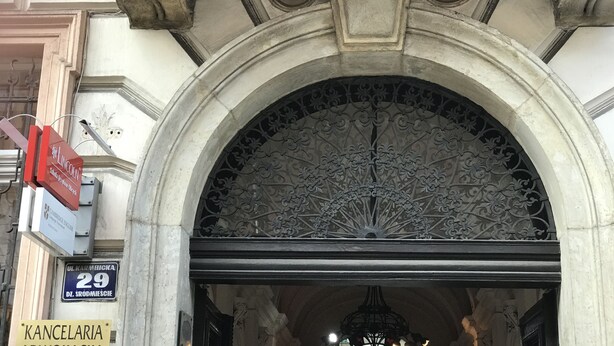 Une entrée architecturale richement décorée, avec un portail en métal et une enseigne indiquant « KANCELARIA ».