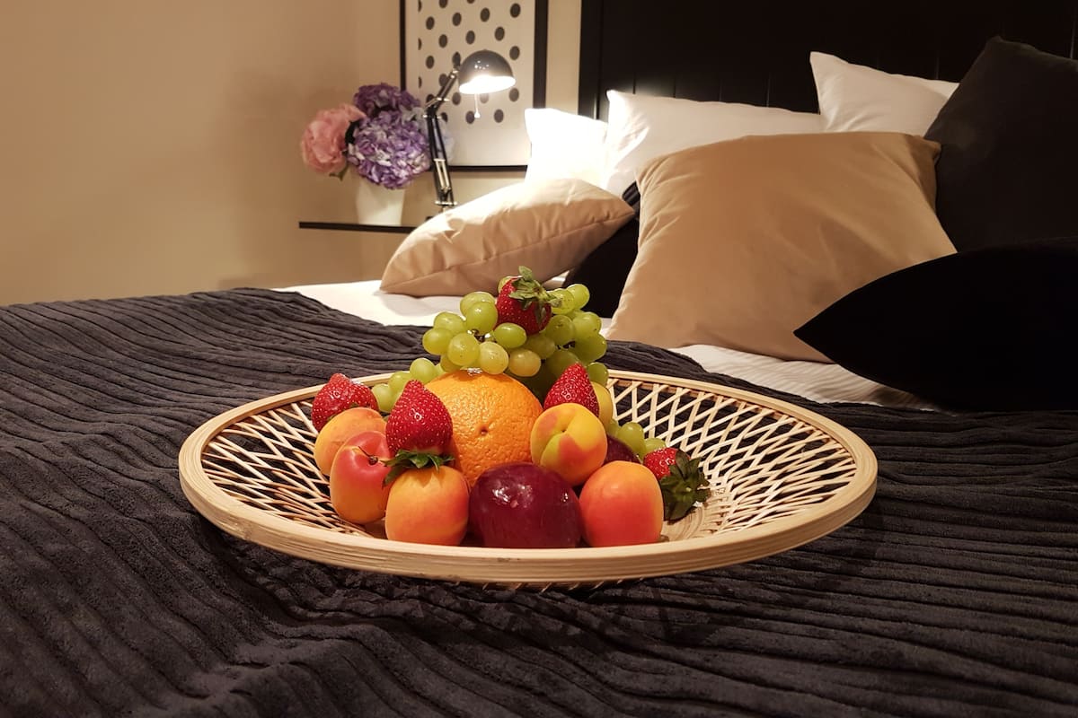 Una cama con una colcha oscura y texturizada, una cesta de mimbre con fresas, uvas, melocotones y una naranja, y una mesita de noche con un jarrón de flores y una lámpara.