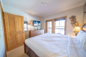 A bedroom with a large bed, wooden wardrobe, a television on a dresser, and a window with blinds.